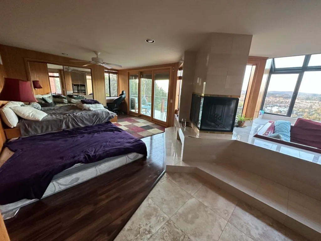 A spacious bedroom with a fireplace, mirrored wall, and windows onto the hills