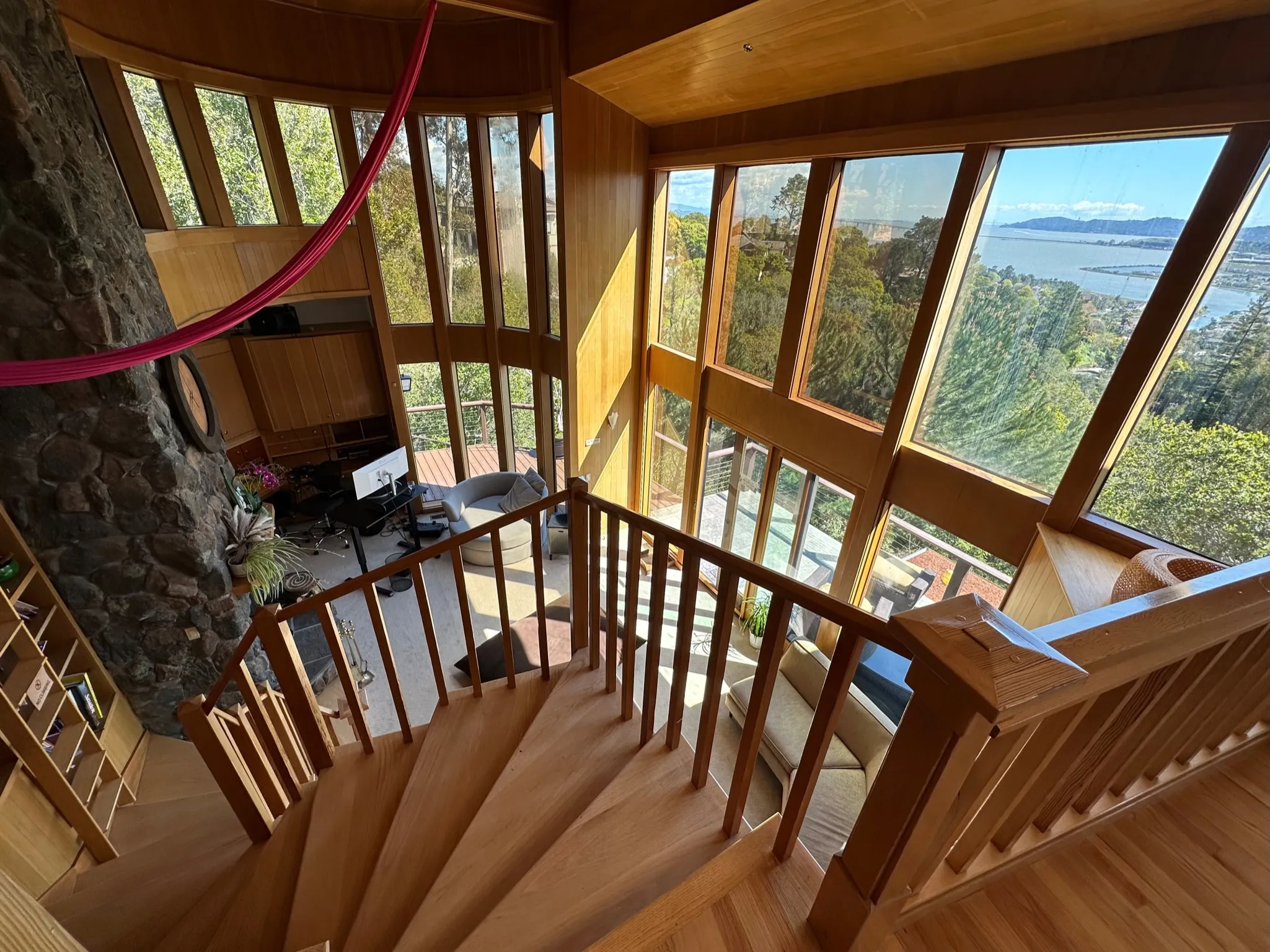 A curved wooden staircase descending past tall windows with a view of the bay